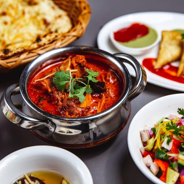 Indian lamb curry in a metal pot, with naan, samosas, and salad on a dark table.
