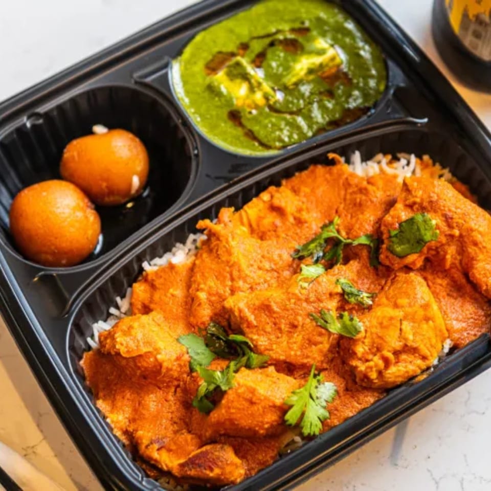 Black takeout tray holding spiced orange chicken over rice, green chutney, two gulab jamuns, and fresh cilantro.