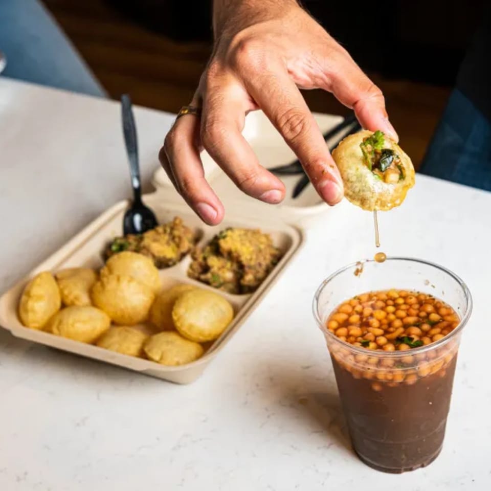 Hand holds a stuffed golden puri over a tray of puris and a glass bowl of chickpea curry.